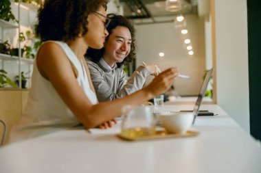 İşbirlikçi meslektaşlar kahve ve dizüstü bilgisayarlarda gülümseyerek telefon ekranındaki verileri analiz ediyorlar.