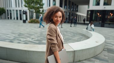 Siyah kadın elinde dizüstü bilgisayarla meydanda yürüyor, kameraya doğru dönüyor, spor ceket giyiyor, kasıtlı adımlar atıyor.,