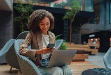 Siyah girişimci parlak lobide dizüstü bilgisayar ve telefonla anlaşma yapıyor, kontrat olarak neşeli bir ifade.