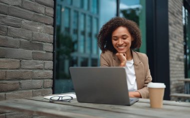 Açık havada bilgisayar, kahve, kitap ve notlarla çalışırken gülümseyen siyah öğrenci.