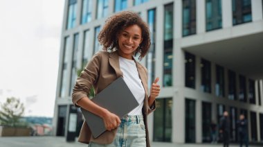 Dizüstü bilgisayarı ve klasörü olan siyahi bir kadın gülümsüyor, hoş bir jest yapıyor, şehir meydanı geçmişi var, sıradan.