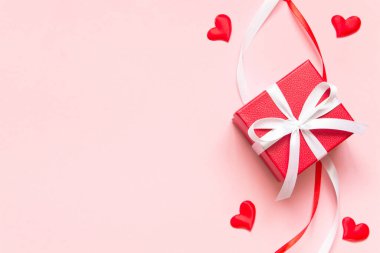 Beyaz kurdeleli kırmızı hediye kutusu, kırmızı ve beyaz dekoratif kurdele ve pembe arka planda birkaç küçük ısıtıcı. Hediye verme kavramı, sevgi, sevgililer günü. Uzayı kopyala, düz olarak