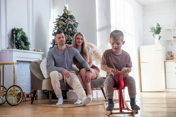 Little son rides a wooden horse while young parents relax at home on the sofa, little boy has fun, family leisure in the living room, friendly and happy family