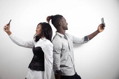 Afrikalı Amerikalı çift, beyaz stüdyo arka planındaki telefonlarda selfie çekiyorlar. Bloglama ve kablosuz teknoloji kavramı.