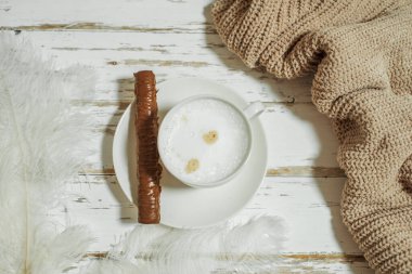 Kadınların elleri süveter içinde, bir fincan kahveyi ahşap masada tutarken. Tabakta çikolata.