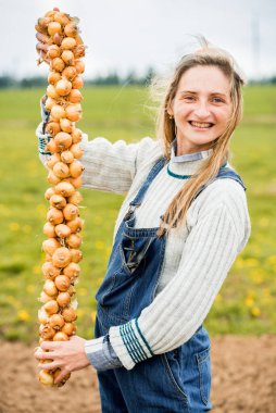 Kot pantolon giymiş gülümseyen kadın çiftçi bahçedeki yeşil soğanın elini tutuyor. Boynunda bir demet soğan var. Ampul dikmek ve hasat yapmak. Sebze buketi
