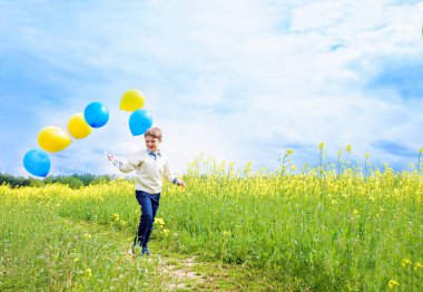 Happy child with balloons running on field.Laughing toddler boy playing in a yellow field. Blooming rapeseed, blue sky with white clouds. Freedom concept
