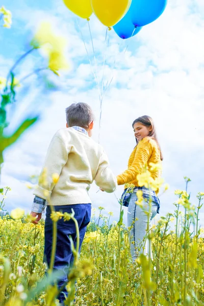 Vertical portrait happy two children run on a yellow field, blooming rapeseed. blue sky and clouds. Blue and yellow balloons, concept of freedom, summer. Holiday and birthday. Central Europe — Fotografia de Stock