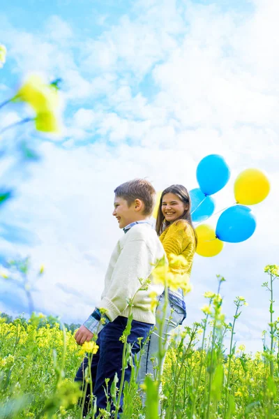 Excited kids with balloons run in field.Jump and fly in blooming yellow rape. blue sky, green grass. Summer beautiful card. Freedom concept. — Stock Photo, Image