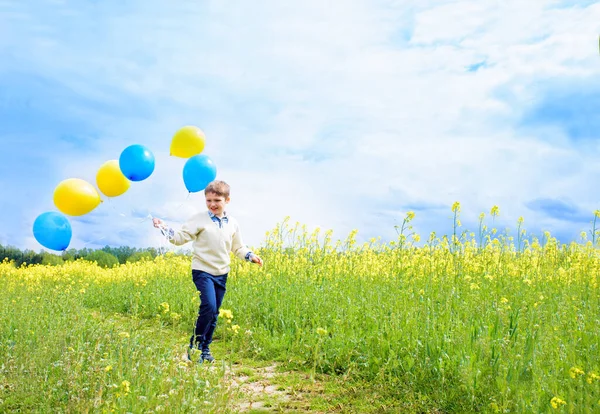 Happy child with balloons running on field.Laughing toddler boy playing in a yellow field. Blooming rapeseed, blue sky with white clouds. Freedom concept — Stock Photo, Image