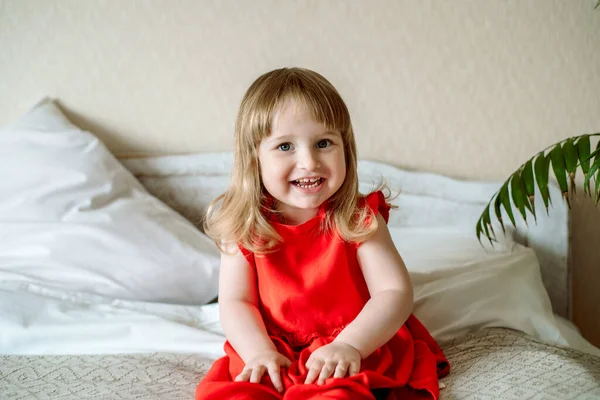 Glad baby porträtt. Känslomässig liten flicka på sängen i ett ljust sovrum. Mysigt vitt rum, glada barn. känslomässigt skriker, skrattar och glädjer.Vit filt, röd klänning. — Stockfoto