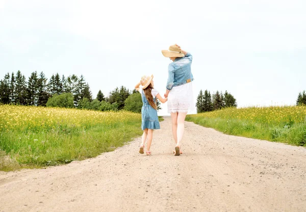 Mother woman little girl my daughter go on rural road through green field in summer.Yellow field, sunset sun, summer day and evening. go into the distance. back — Stock Photo, Image
