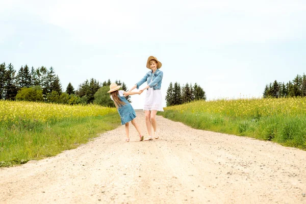 Mother woman little girl my daughter go on rural road through green field in summer.Yellow field, sunset sun, summer day and evening. go into the distance. swirl, rejoice, active life position. — Stock Photo, Image
