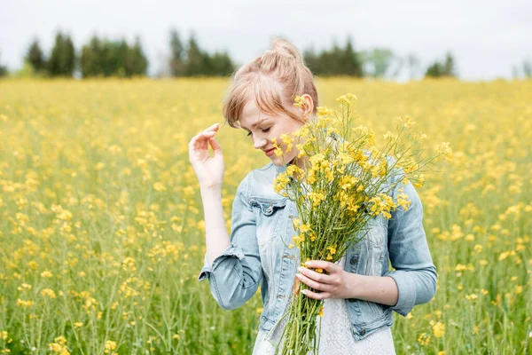 Lovely blonde girl in a rapeseed flowering field, tender and embarrassed. yellow flowers, a bouquet in her hands. Summer day in the village. Freedom, air is the wind — Stock Photo, Image