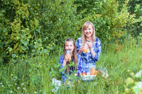 Outdoors park picnic. Beautiful young mom and little girl sitting in forest and enjoying.white bread, pastries, fruits in basket, apples and bananas.Drink orange juice from glass from straw. mustache. — Stock Photo, Image