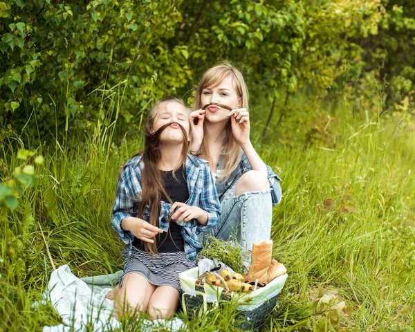 Mãe e filha estão brincando e se divertindo juntos. Menina bonita e mãe têm bigode. Piquenique na floresta. parque de verão, cesta de frutas, pão branco. Ao ar livre. Férias em família e união. — Fotografia de Stock