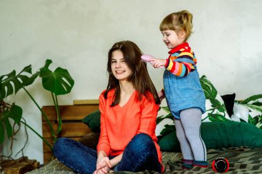 Tatlı küçük çocuk aynanın yakınını tarıyor. Mutlu kız evde, yatağın üzerinde çocuk odasında eğleniyor. Egzotik bitkiler. Yeşil ve gri ekose. Hemşire bakımı. Aile sevgisi kavramı.