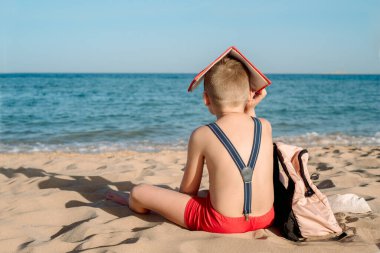 Mutlu genç çocuk güneşli bir günde kitap okuyor. Çocuk sahilde sırtı dönük oturuyor. Tatilin son günü. Okula dönelim. Sırt çantası ve pantolon askısı, kırmızı defter. Kötü ruh hali ve öğrenme arzusu yok..