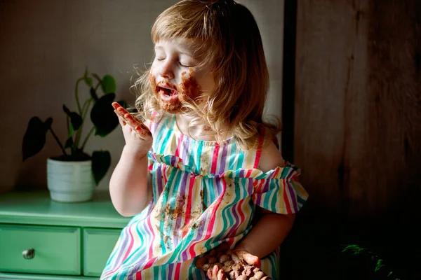 可爱的卷发女婴吃巧克力，孩子涂了蜂蜜。肮脏的脸颊、鼻子和双手。7月7日巧克力庆祝会对孩子有害的糖果。不健康的食物室内植物. — 图库照片