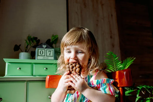 Bonito bebê encaracolado menina comendo chocolate. A criança é alérgica à menina doce e suja, o dia 7 de julho é dia de chocolate. Casas, plantas indoor. Foco seletivo — Fotografia de Stock