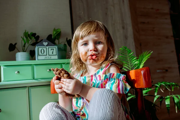 Bonito bebê encaracolado menina comendo chocolate. A criança é alérgica à menina doce e suja, o dia 7 de julho é dia de chocolate. Casas, plantas de interior. — Fotografia de Stock