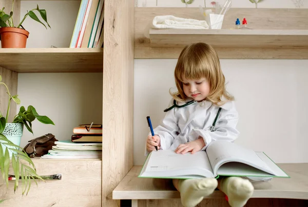 Menina do bebê uniformizado como médico escrevendo para clipboard.Medical vestido e fonendoscópio. O médico da enfermaria escreve um histórico médico. — Fotografia de Stock