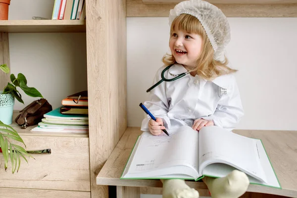 Cute little baby girl in doctor uniform with clipboard in hospital.Phonendoscope and white bathrobe. Notepad and history of disease. The concept of health, medicine and laboratory diagnostics. — Stock Photo, Image
