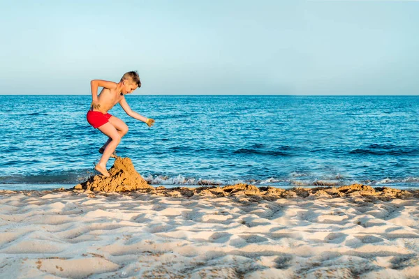 Wakacje na morzu. Słodki chłopiec na plaży nad morzem bawi się piaskiem. Rzuca zamek z piasku i ciasta. zachód słońca, błękitne niebo i relaks koncepcji podróży. przestrzeń kopiowania — Zdjęcie stockowe