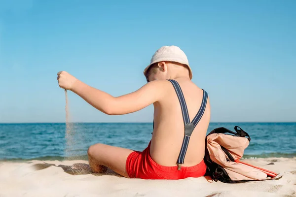 Λυπηρό αγόρι που διαβάζει ένα βιβλίο την ηλιόλουστη μέρα. αγόρι που κάθεται με την πλάτη του στην παραλία. Τελευταία μέρα διακοπών. Πίσω στο σχολείο. Σακίδιο και τιράντες, κόκκινο βιβλίο. Κακή διάθεση και όχι επιθυμία για μάθηση.. — Φωτογραφία Αρχείου