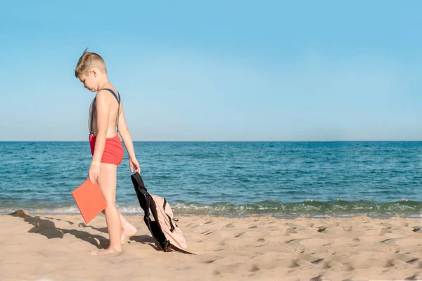 Glücklicher kleiner Junge, der an einem sonnigen Tag ein Buch liest. Junge, der mit dem Rücken am Strand sitzt. Letzter Urlaubstag. Zurück zur Schule. Rucksack und Hosenträger, rotes Buch. Schlechte Laune und kein Lernwille. — Stockfoto