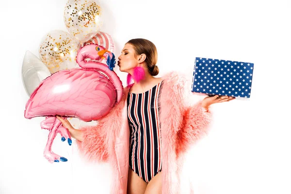 Emotional girl at a party in a red striped swimsuit and a pink fluffy fur coat with balloons. Kisses a pink flamingo. Joyful woman woman holding a lot of boxes with gifts on a white background