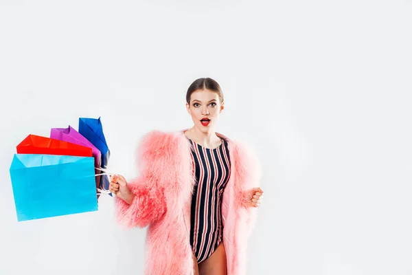 Young fashionable sexy pretty girl in pink coat of fur with hood and fashion makeup holding colorful package in studio.in swimsuit is skipping for shopping, a lot of bright multi colored packages — Stock Photo, Image