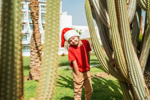 Sad boy in santa costume on the beach. In a red hat santa claus. Christmas Holidays. sea vacation. blue sky, palm trees and tall cacti of Egypt or Turkey. Plays in the hotel. Stock Picture