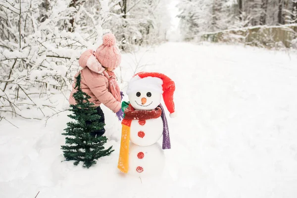 Счастливый ребенок играет со снеговиком. Смешная маленькая девочка на прогулке зимой на открытом воздухе — стоковое фото