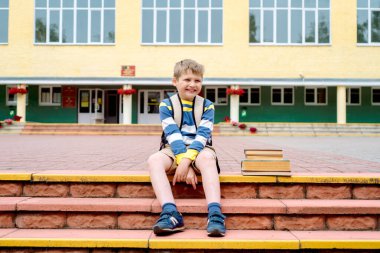 Güzel okul çocuğunun portresi gündüz vakti dışarıda çok mutlu görünüyor. Basamaklarda kitaplar ve büyük bir okul çantasıyla oturmak. Üzgün ve mutsuz duygusal bebek.