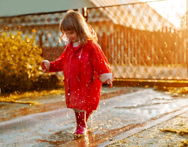 Linda chica con una chaqueta roja está saltando en el charco.La puesta de sol cálido verano u otoño. verano en el pueblo. — Foto de Stock