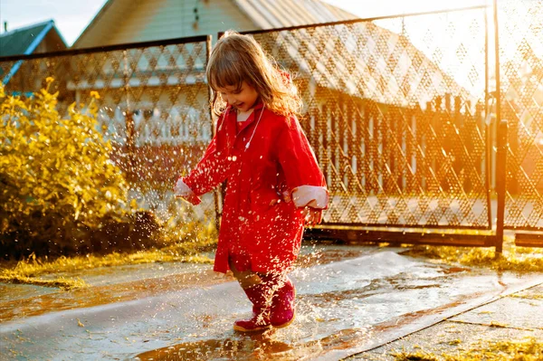 Linda chica con una chaqueta roja está saltando en el charco.La puesta de sol cálido verano u otoño. verano en el pueblo. — Foto de Stock