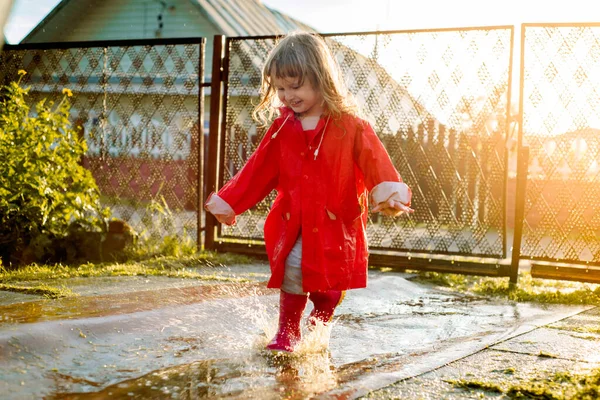 Menina bonito em uma jaqueta vermelha está pulando no pudim. O pôr do sol quente de verão ou outono. verão na aldeia. — Fotografia de Stock