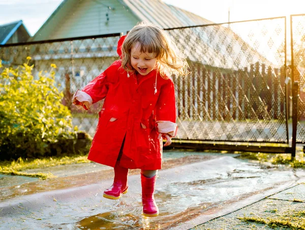 Linda chica con una chaqueta roja está saltando en el charco.La puesta de sol cálido verano u otoño. verano en el pueblo. — Foto de Stock
