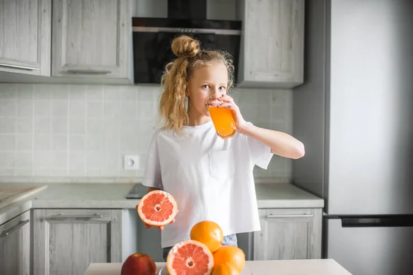 Niña caucásica bebiendo jugo de naranja en el interior sentado en la mesa de la cocina. Concepto de nutrición alimentaria saludable para niños. — Foto de Stock