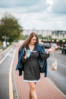 Elinde kahve ve telefon olan bir kız, işe gitmek için sokakta yürüyor. Genç kadın işçi yaşam tarzı.