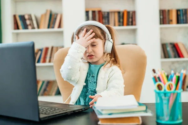 Emotional Caucasian little four years old girl doing homework using the notebook for distance learning sitting at the table. Distance online learning concept. Stock Picture