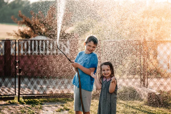 Bambini piccoli che giocano insieme con un tubo da giardino nella calda e soleggiata giornata estiva al tramonto — Foto Stock