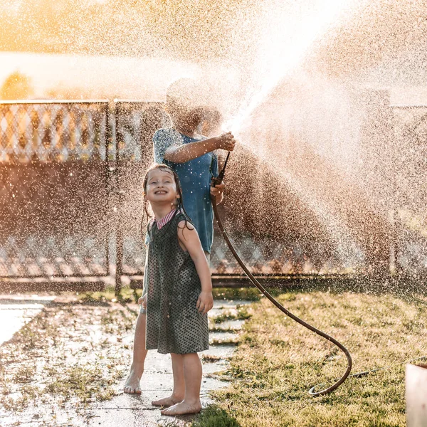 Crianças brincando junto com uma mangueira de jardim no dia de verão quente e ensolarado no pôr do sol — Fotografia de Stock