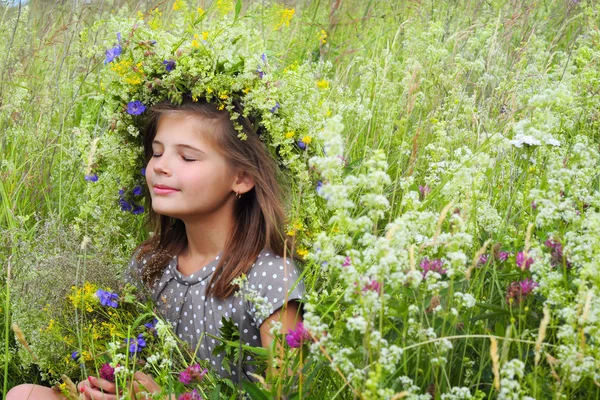 O fată zâmbitoare fericită, cu ochii închişi pe o pajişte. O coroană florală pe cap — Fotografie, imagine de stoc