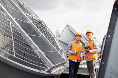 Asyalı erkek mühendis boru tesisatlarını ve soğutucuları inceliyor. Fabrikadaki soğutma tesisi. Kask takan erkek tamirci ve yansıtıcı güvenlik yeleği kondisyon sistemlerinde dizüstü bilgisayar, tablet ve soğutma kuleleri olan ekipman kullanıyor.