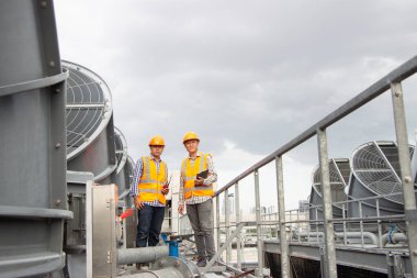 Asyalı erkek mühendis boru tesisatlarını ve soğutucuları inceliyor. Fabrikadaki soğutma tesisi. Kask takan erkek tamirci ve yansıtıcı güvenlik yeleği kondisyon sistemlerinde dizüstü bilgisayar, tablet ve soğutma kuleleri olan ekipman kullanıyor.