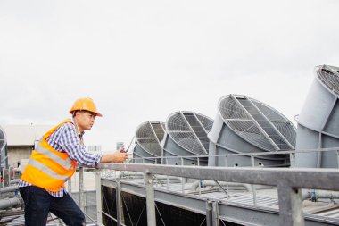 Asyalı erkek mühendis boru tesisatlarını ve soğutucuları inceliyor. Fabrikadaki soğutma tesisi. Kask takan erkek tamirci ve yansıtıcı güvenlik yeleği kondisyon sistemlerinde dizüstü bilgisayar, tablet ve soğutma kuleleri olan ekipman kullanıyor.