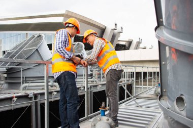 Asyalı erkek mühendis boru tesisatlarını ve soğutucuları inceliyor. Fabrikadaki soğutma tesisi. Kask takan erkek tamirci ve yansıtıcı güvenlik yeleği kondisyon sistemlerinde dizüstü bilgisayar, tablet ve soğutma kuleleri olan ekipman kullanıyor.