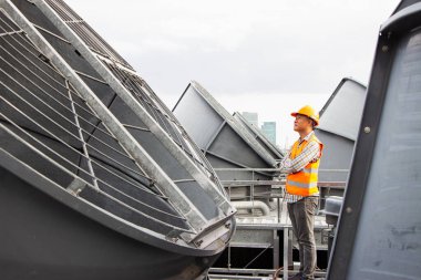 Asyalı erkek mühendis boru tesisatlarını ve soğutucuları inceliyor. Fabrikadaki soğutma tesisi. Kask takan erkek tamirci ve yansıtıcı güvenlik yeleği kondisyon sistemlerinde dizüstü bilgisayar, tablet ve soğutma kuleleri olan ekipman kullanıyor.